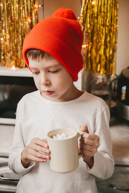 Boy in a white t-shirt and a red hat drinking cocoa with marshmallows in the kitchen. Christmas mood. Christmas sweets and drinks