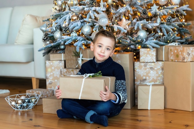 A boy with a Christmas gift in his hands sitting under a Christmas tree and looks into the camera. There are many wrapped Christmas gifts under the tree. Cozy winter morning at home.