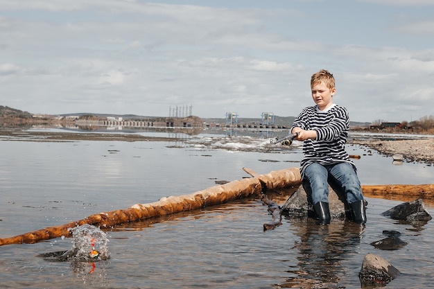 A boy with a fishing rod is fishing, sitting on the bank of the river. A handsome boy in a striped vest sits with a fishing rod in his hands