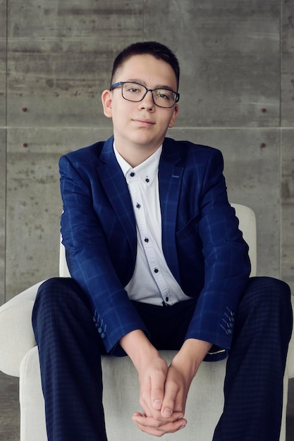 A boy with glasses with an intelligent look sits in a chair with his hands clasped in a lock