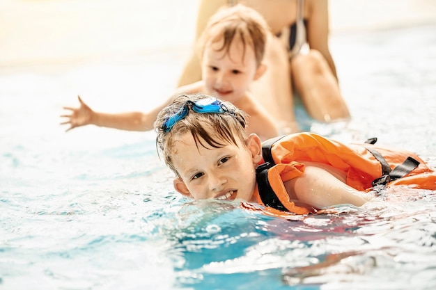 Boy with googles and safety vest learns to swim near brother and mom in large pool in aquapark