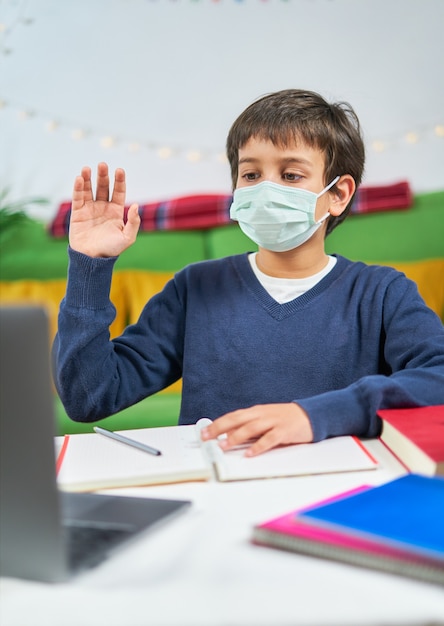 Boy with mask having a video conference on laptop with teacher from home, wearing headphones and waving on screen, free space