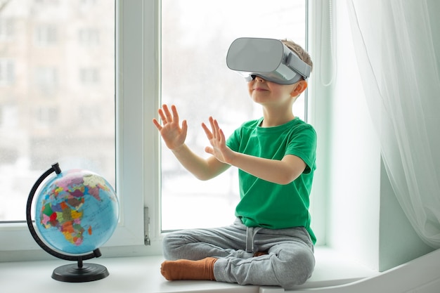 A boy with a virtual reality headset is sitting at home at the table indoors The child explores the world of virtual reality through video and games