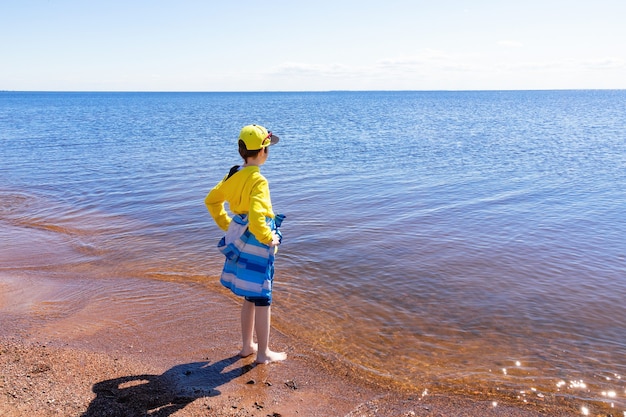 Boy in yellow hoodie and cap, in blue jacket, with rolled up jeans barefoot on the seashore on a sunny day.