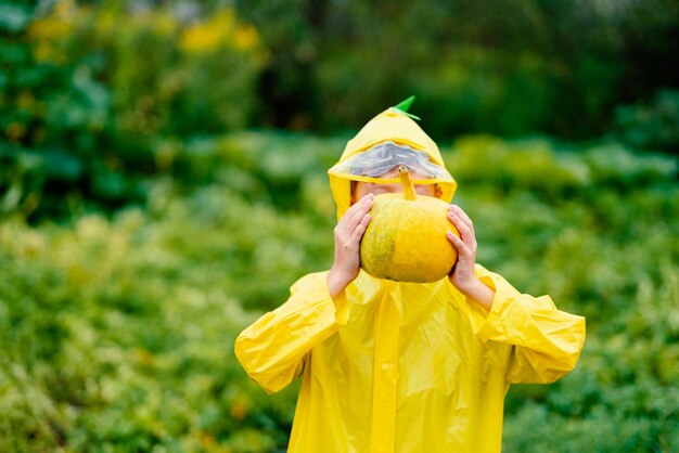 Boy in a yellow raincoat holds a yellow pumpkin Preparing for Halloween Pumpkin harvest Pumpkin in the hands of a boy near the face against a background of greenery