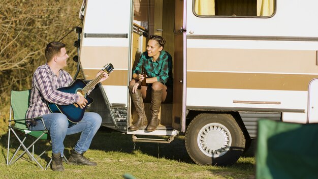 Boyfriend in front of the retro camper van singing on guitar for his beautiful girlfriend. Cheerful couple in wilderness with camper.