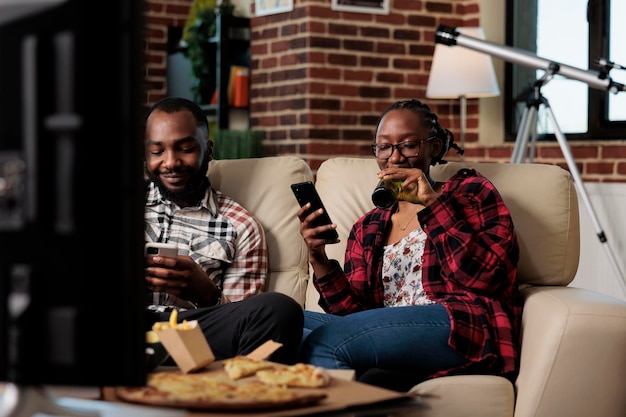 Boyfriend and girlfriend using smartphones and eating takeaway food while they watch movie on television. Takeout pizza, noodles, burger and snacks with bottles of beer watching tv film.