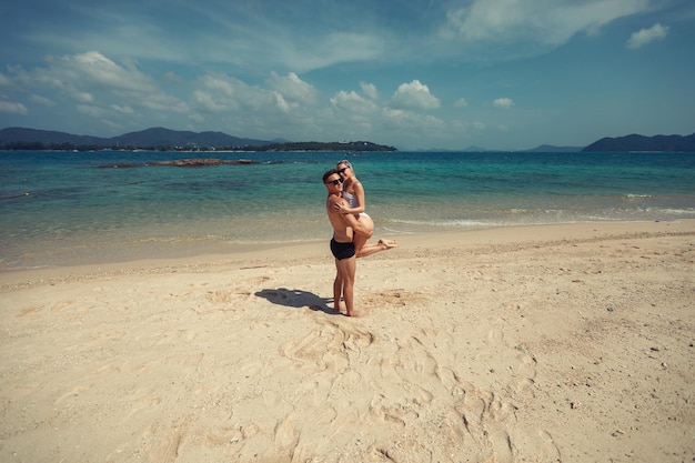 Boyfriend holds his slim girlfriend with long cornrows and white swimsuit in his arms, sand and picturesque sky; love concept.