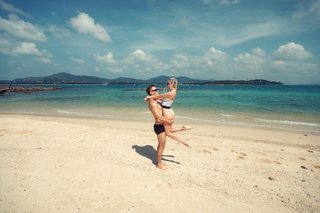 Boyfriend holds his slim girlfriend with long cornrows and white swimsuit in his arms, sand and picturesque sky; love concept.