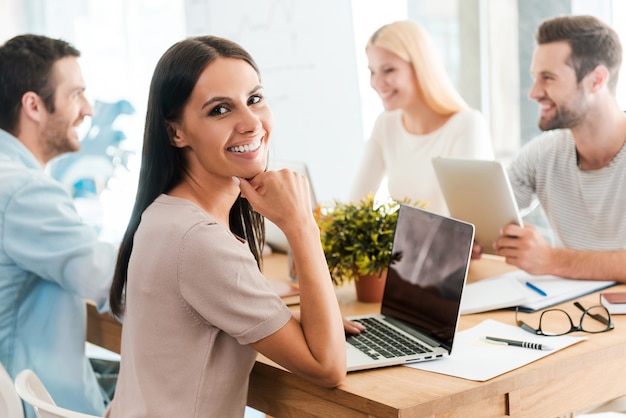 Brainstorming with colleagues. Beautiful young woman holding hand on chin and smiling while sitting together with her colleagues at the desk in office