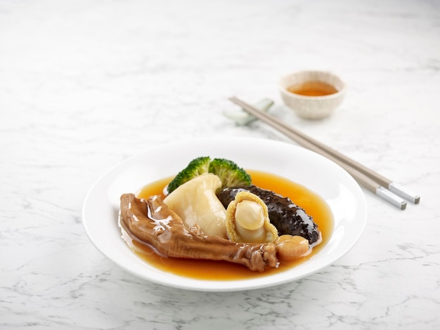 Braised Sliced Abalone with Conpoy Goose Web Sea Cucumber and Fish Maw in Brown Sauce served in a dish side view on grey background