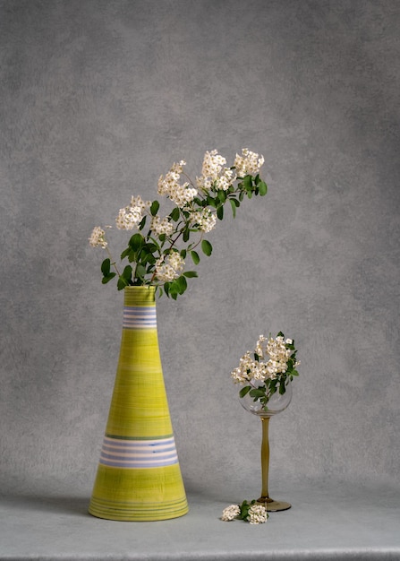 A branch with white flowers in a green clay vase on a gray background In a glass goblet is a small