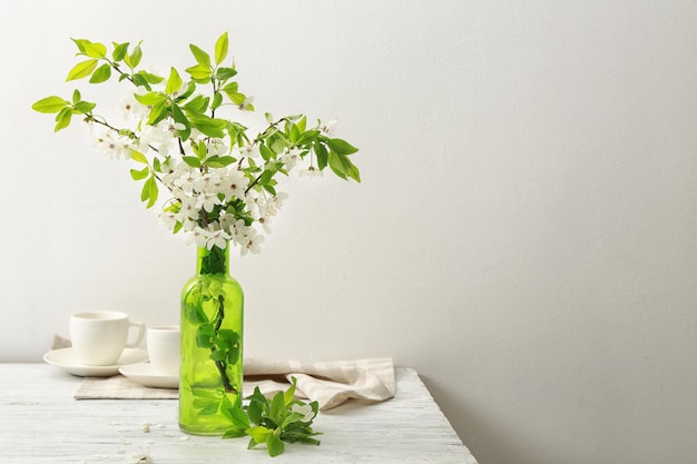 Branches of blooming tree flowers and leaves in glass vase on wooden table with cups