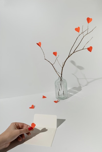 Photo branches with red paper hearts and female hand putting heart in white envelope minimal love or valentine concept