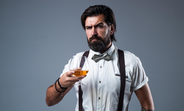 Brandy or cognac bearded man in suspenders drink scotch whiskey brutal guy bartender wear bow tie elegant male barman handsome hipster drinking rum glass party goer with alcohol