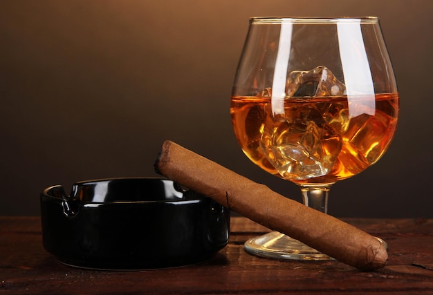 Brandy glass with ice and cigar on wooden table on brown background