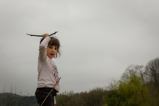 Brave little girl holding a stick with hand up at park on cloudy day