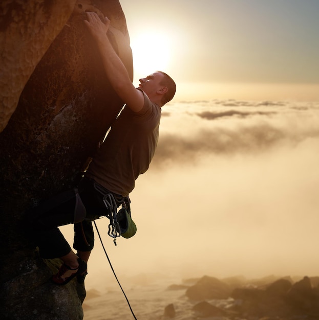 Brave male climber blinded by sunlight on high natural rock wall over gray clouds Climbing with rope and safety harness