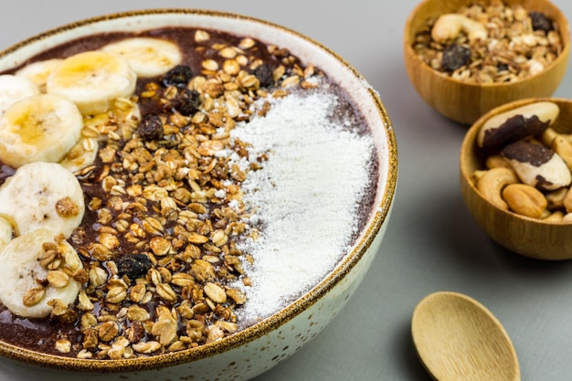 Brazilian acai in a white bowl with banana powdered milk honey granola and nuts in small bamboo bowls Cashew nuts and Para Grey background Selective focus