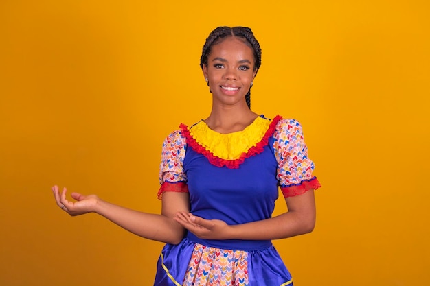 Brazilian afro woman wearing typical clothes for the Festa Junina in yellow background pointing with copy space