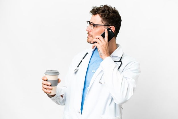 Brazilian doctor man over isolated white background holding coffee to take away and a mobile