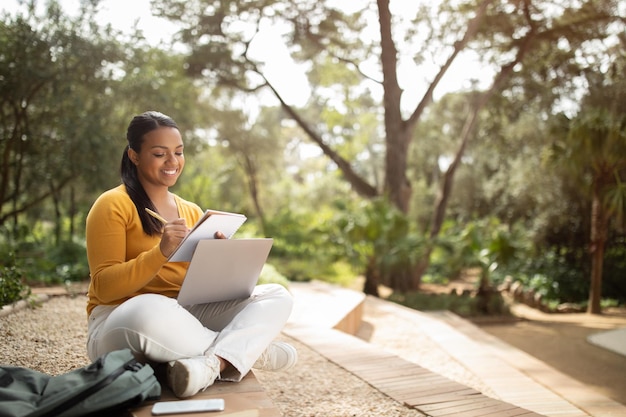 Brazilian lady student writing something in notebook sitting with laptop in park and studying