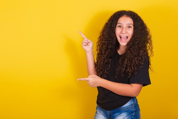 Brazilian latin american girl for afro hair yellow background smiling suggesting suggestion indication pointing with hands to the side negative space for advertising and publicity text recommending