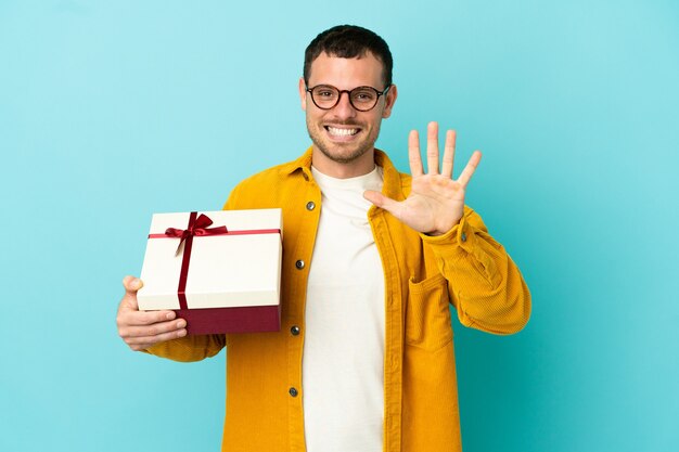 Brazilian man holding a gift over isolated blue background counting five with fingers