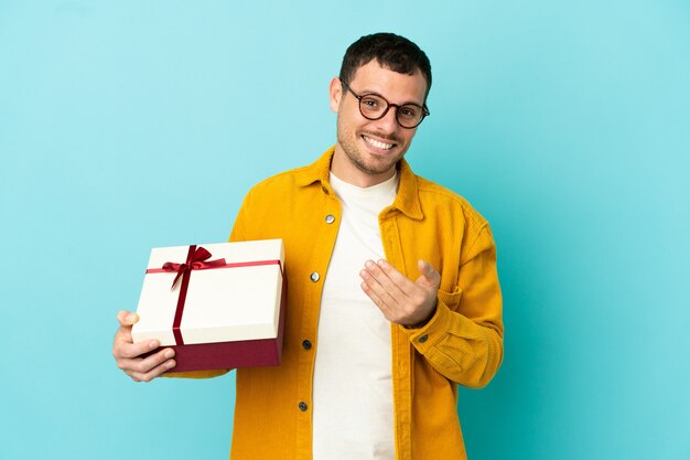 Brazilian man holding a gift over isolated blue background inviting to come with hand. Happy that you came