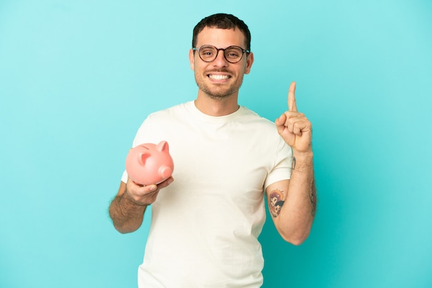 Brazilian man holding a piggybank over isolated blue background showing and lifting a finger in sign of the best