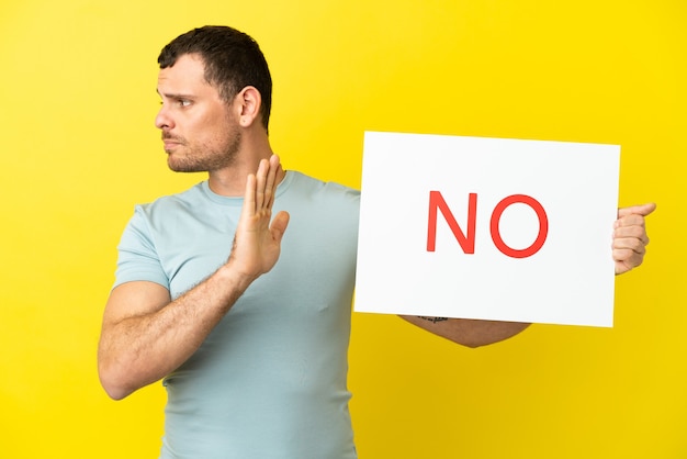 Brazilian man over isolated purple background holding a placard with text NO and doing stop sign