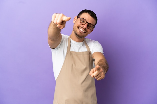 Brazilian restaurant waiter over isolated purple background points finger at you while smiling