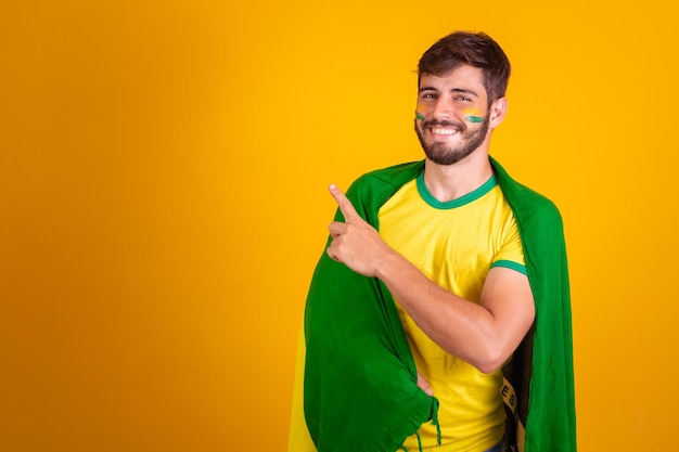 Brazilian supporter with a brazil flag on his back on yellow background pointing to space for text