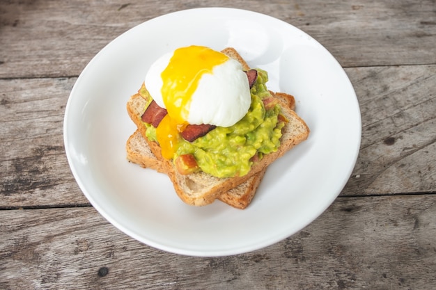 Bread, avocado sauce, bacon, spinach, egg benedict on white plate and old wood background