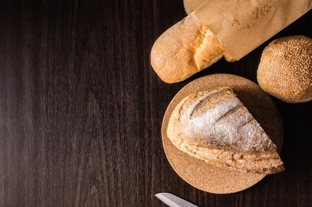 Bread concept a half loaf of bread on the wooden plate, a loaf of bread into a paper bag, a knife, and a bun put on a black wooden scene.