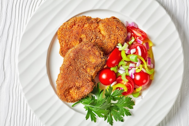 Breaded lamb chops served with tomato, red onion, green pepper salad on a white plate on a wooden table, flat lay, close-up, macro