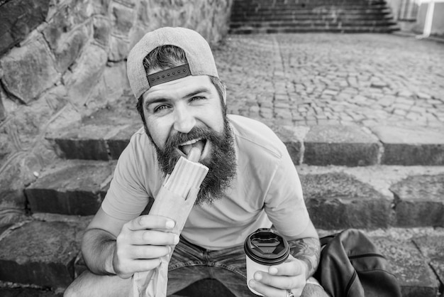 Break for sausage Caucasian guy having meal break on urban background Bearded man having hot dog and coffee for snack break Hipster snacking on stairs during rest break