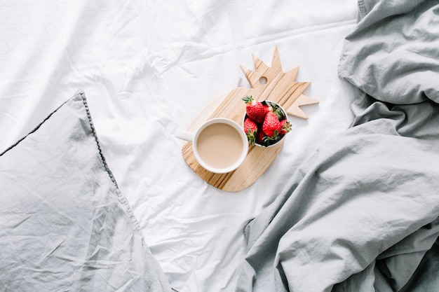 Breakfast in bed with coffee mug and strawberry. Bright grey linens. Flat lay, top view.