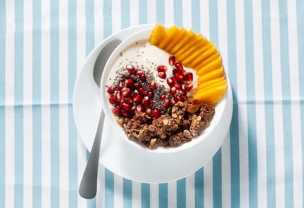 Breakfast bowl of granola, yogurt, fresh sweet mango, chia seeds, pomegranate seeds and maple syrup on a blue striped napkin