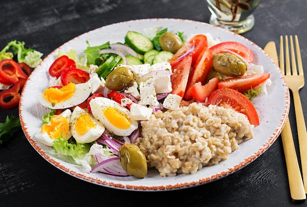 Breakfast oatmeal porridge with greek salad of tomatoes, cucumbers, olives, feta cheese and eggs. Healthy balanced food.