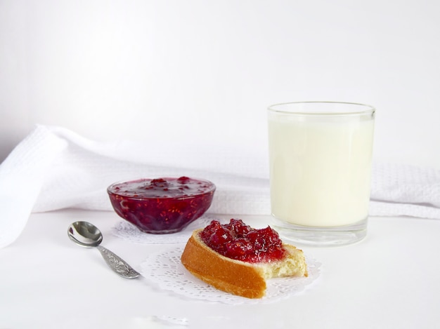 Breakfast one glass of milk with a nibbled piece of bread with raspberry jam, as well as a bowl of jam on a white surface