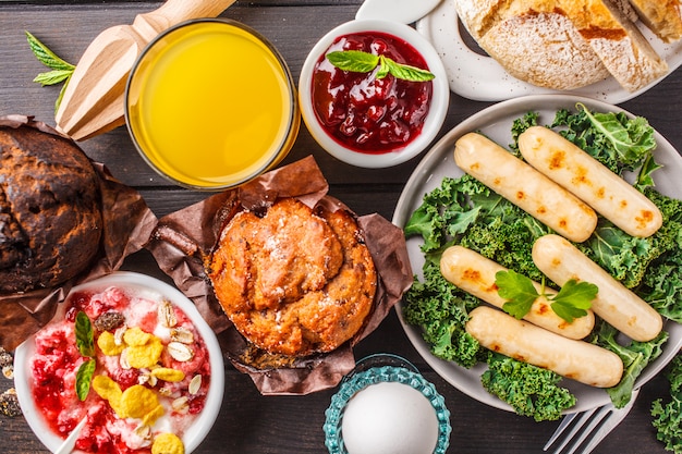 Breakfast served with muffins, grilled sausages, juice, fresh bread and parfait on a dark wooden table.