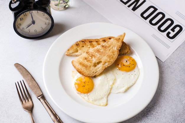 Breakfast time. Two fried eggs with toast for breakfast on a textured light gray background with a newspaper and an alarm clock