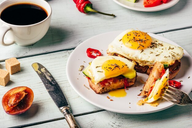 Breakfast toasts with vegetables and fried egg on white plate, cup of coffee and some fruits over wooden