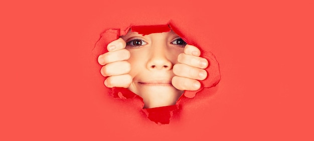 Breaking paper background Emotions concept Positive child with toothy pleasant smile on face keeps through torn hole in red paper