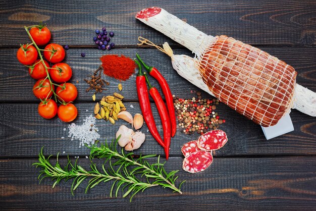 Bresaola and sliced cured sausage with spices, chili pepper, garlic and tomatoes on dark wooden rustic background.