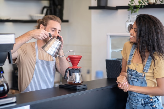 Brewing method. Happy male barista standing behind bar making coffee with pourover and watching long haired woman