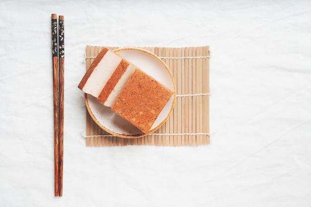 Brick of organic somoked tofu cheese served on white plate over a bamboo mat. Flat lay. Top View. Vegan snack concept