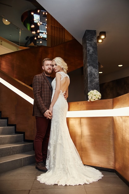 Bride in a chic long dress with a train and the groom stand on big stairs, couple in love hugs on stairs kiss and look at each other. Day of the wedding, couple on the day of registration of marriage