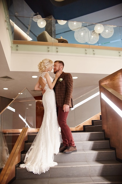 Bride in a chic long dress with a train and the groom stand on big stairs, couple in love hugs on stairs kiss and look at each other